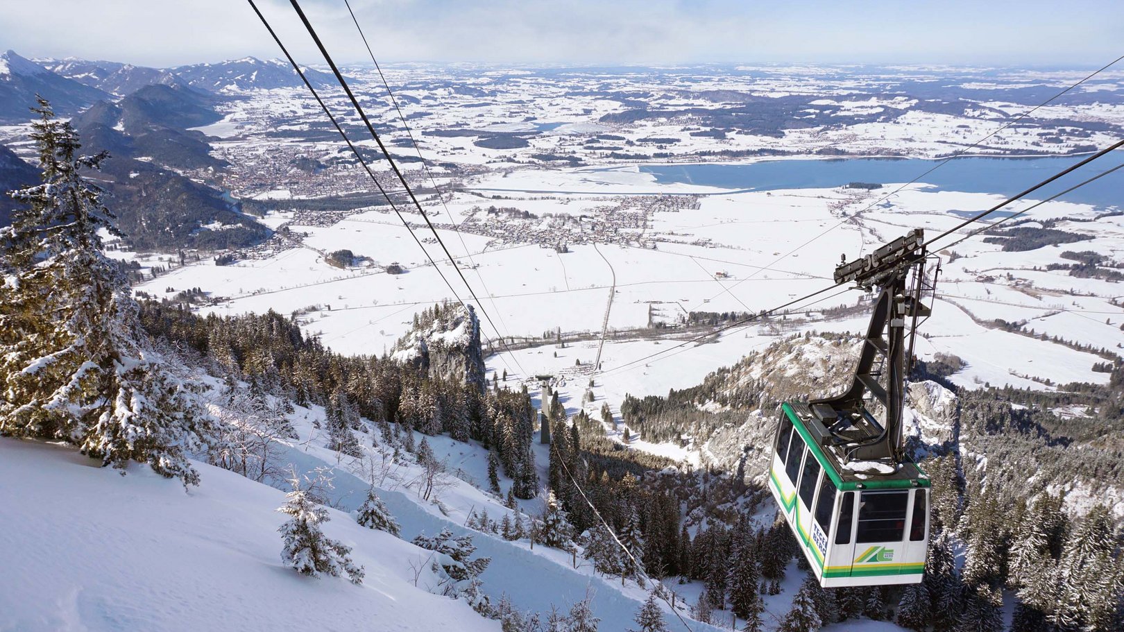 Skifahren im Allgäu | Hotel Sommer Füssen Seilbahn fährt über verschneite Berge mit Tal und See im Hintergrund