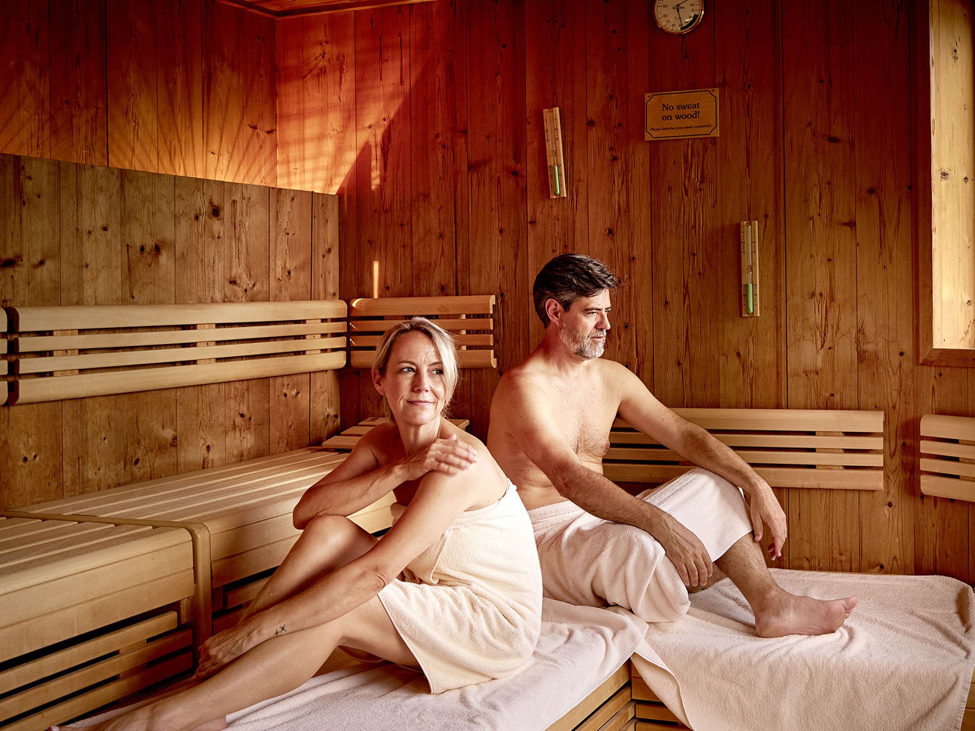 Wellnesshotel in Füssen im Allgäu | Hotel Sommer Paar entspannt in Holzsauna mit Handtüchern und Fensterblick
