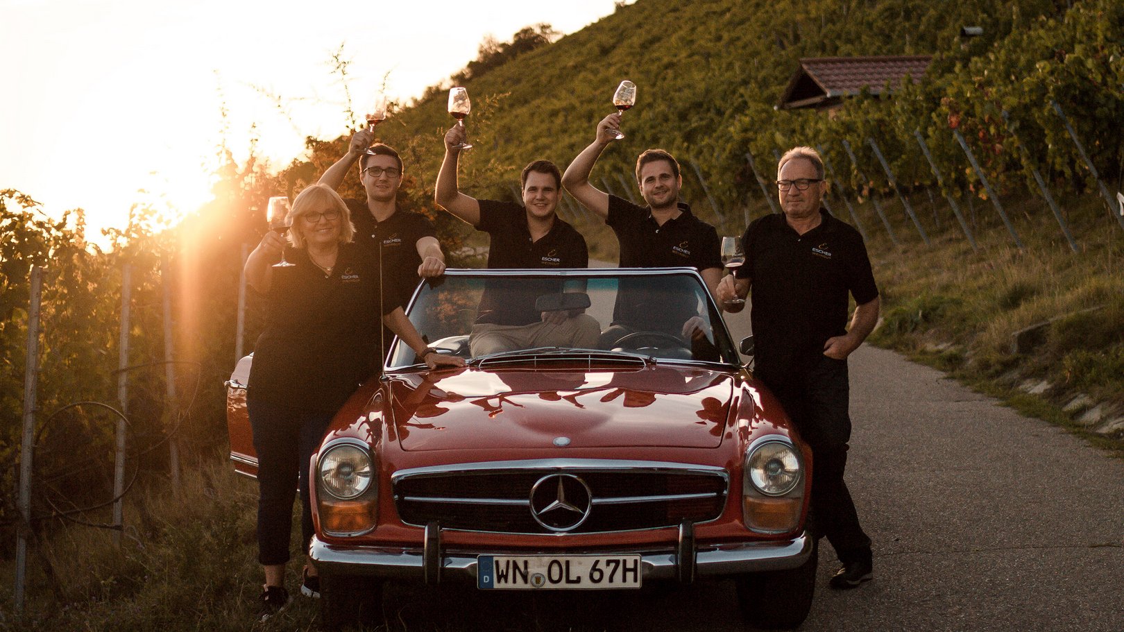 Kulinarikwochen – das Jahresprogramm Fünf Personen mit Weingläsern neben rotem Oldtimer im Weinberg bei Sonnenuntergang